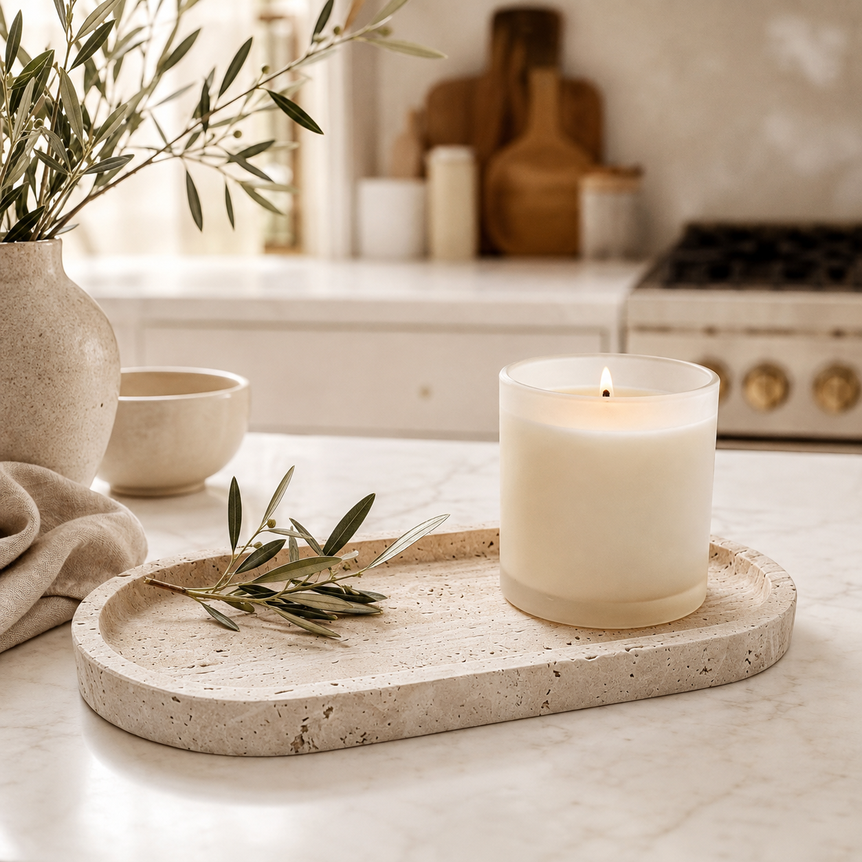 Oval travertine tray styled with a white candle and olive branches on a warm neutral kitchen countertop.