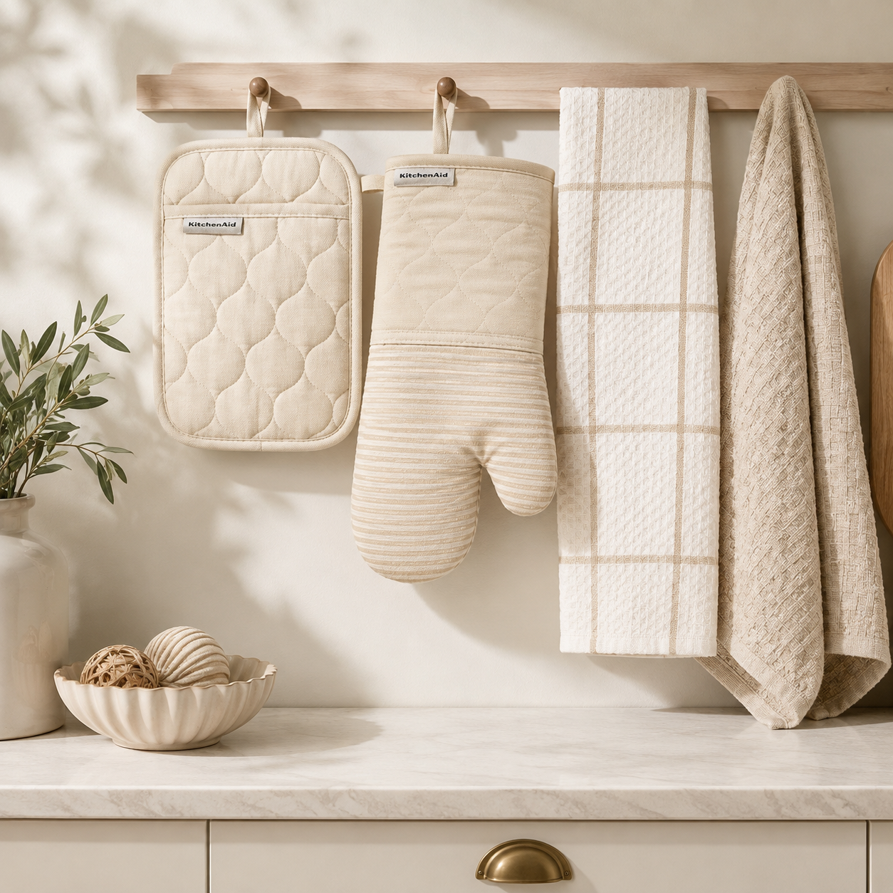 Quilted pot holder, oven mitt, and two patterned kitchen towels hanging from a wooden peg rack above a counter.